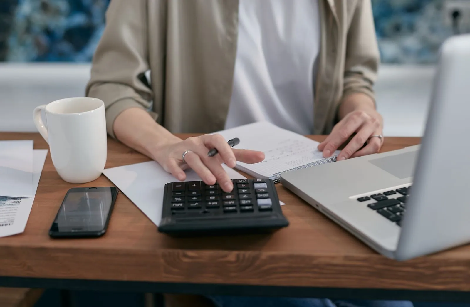 A person using a calculator and laptop while taking notes at a wooden desk.